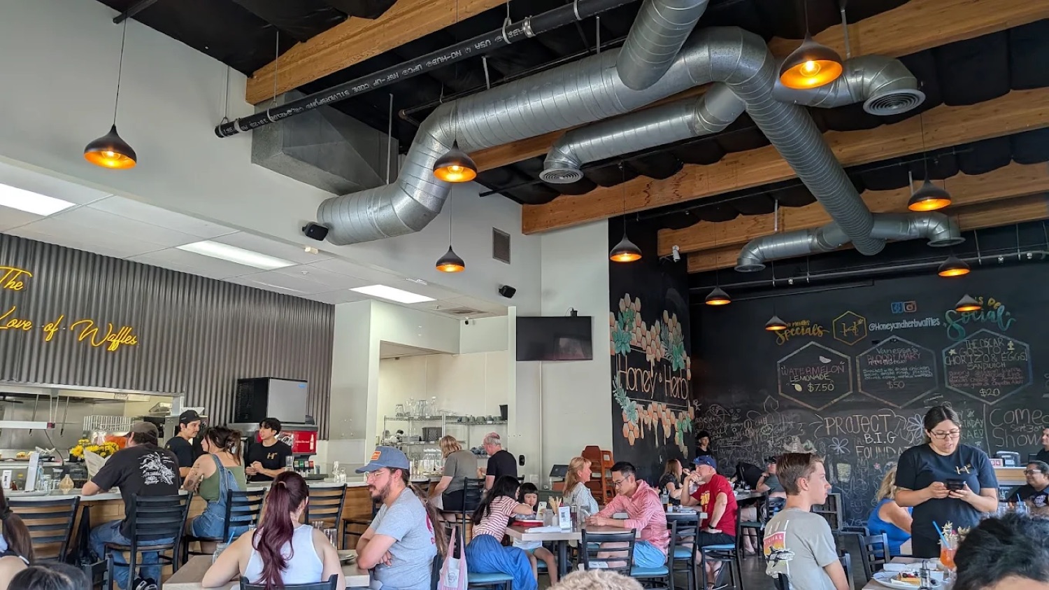 Busy dining room at Honey & Herb Belgian Waffle House with 'The Love of Waffles' neon sign and chalkboard mural